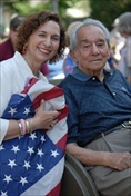 A young woman sitting, smiling and holding an American flag next to an elderly gentleman - also seated and smiling