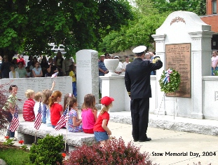 Large marble stone with memorial plaque surrounded by los marble walls. Children are sitting between the 2 walls and in front of memorial stone.  Uniformed officer is facing stone and saluting