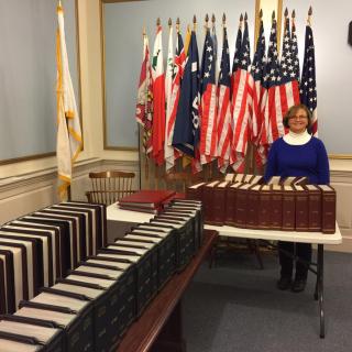 Town clerk standing at a table with volumes of books with several flags behind her