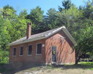 Small brick school house with a chimney, four windows and one door with trees behind it